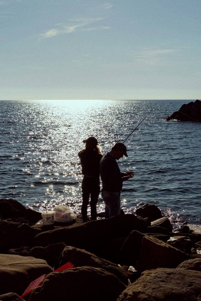 Two anglers on dark rocks with the sun on the sea